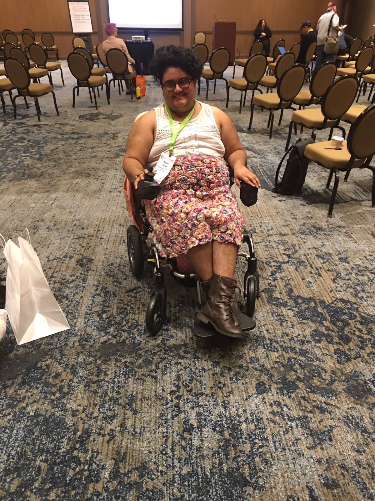 Noor sits in a travel power chair in brown boots and a floral skirt and lace top